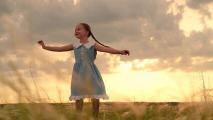 Happy child girl plays spinning in park on grass. Child girl in dress dances spinning with her arms out to sides. Child dream to dance. Little girl smiles in front of sky. Children's dreams in outdoor
