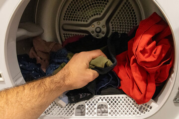Cropped view of a person removing clothes from a dryer, showing forearm and mixed laundry inside a...