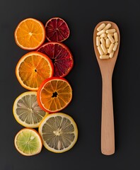 Top view of a wooden spoon filled with vitamin capsules next to sliced citrus fruits like orange and lemon on a black background for immunity and natural supplement concepts.