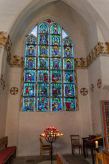 Colorful stained-glass windows depicting biblical scenes adorn a chapel, with candlesticks and simple wooden pews in front.