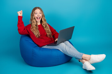 Adorable young woman enjoying laptop usage on a blue beanbag while expressing joy in a casual...