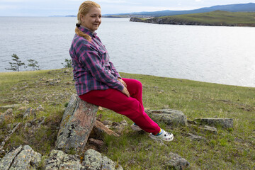 A young white woman sits on a rock near Lake Baikal. She is wearing a plaid shirt and red trousers.