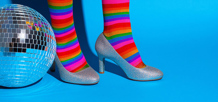 Glitter high heels with vibrant rainbow socks beside a disco ball on a bright blue background, perfect for fashion branding and bold creative campaigns