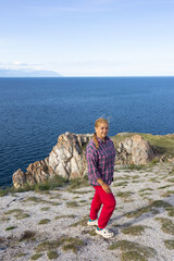 A young white woman sits on a rock near Lake Baikal. She is wearing a plaid shirt and red trousers.