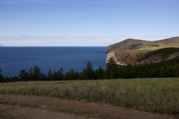 A serene view of Lake Baikal with calm waters reflecting the sky. Mountains and an island are visible in the distance under a cloudy sky.