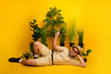 Relaxed man in safari outfit lounges among plants on yellow studio background conveying travel...