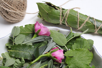 Close-up of fresh lavender, sage, and lemon balm leaves with two roses and a freshly made smudge stick. A roll of natural twine in the background adds a rustic botanical touch.	