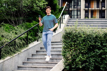 Smiling young man walking down outdoor stairs in urban park during sunny day, holding smartphone, enjoying the moment