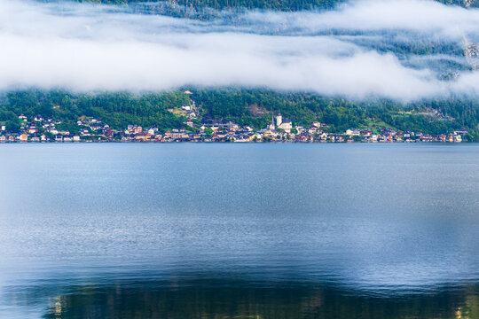 Blick &uuml;ber den stillen See auf Hallstatt, das teilweise im Nebel liegt. Die bunten H&auml;user und die Kirche spiegeln sich im ruhigen Wasser wider.