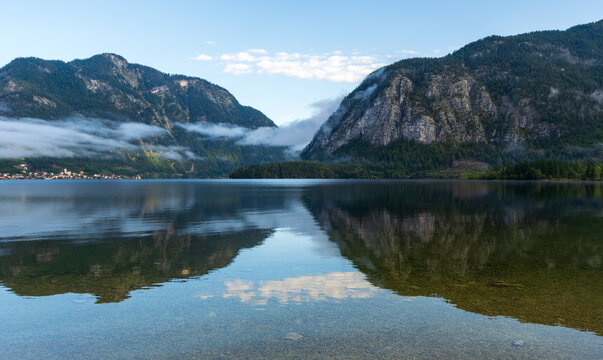 Ein klarer Bergsee spiegelt die umliegenden Berge und Wolken im ruhigen Wasser. Nebelschwaden liegen &uuml;ber dem Tal und verst&auml;rken die friedliche Stimmung.