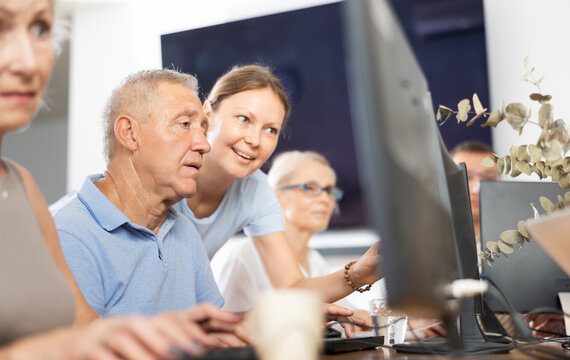 Adult woman teacher shows group of elderly people how to work with computer at computer course