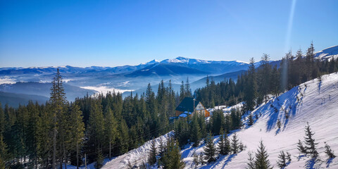 Beautiful winter scene with snow covered pine forest, cozy mountain cabin and glowing sunset over misty hills.