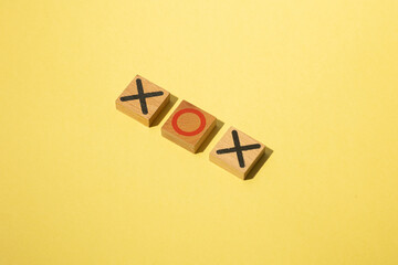 Minimalist arrangement of three wooden tic-tac-toe pieces placed on a yellow background. The pieces display a sequence of X, O, and X, creating a clean and playful visual composition.