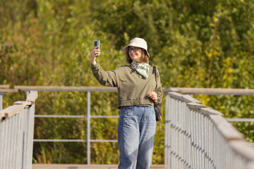 Woman smiling and taking a selfie on a wooden bridge surrounded by lush greenery on a sunny day