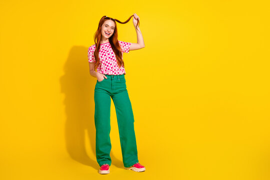 Young girl in pink checkered tee and green pants smiles and plays with her hair against a bright yellow background - Powered by Adobe