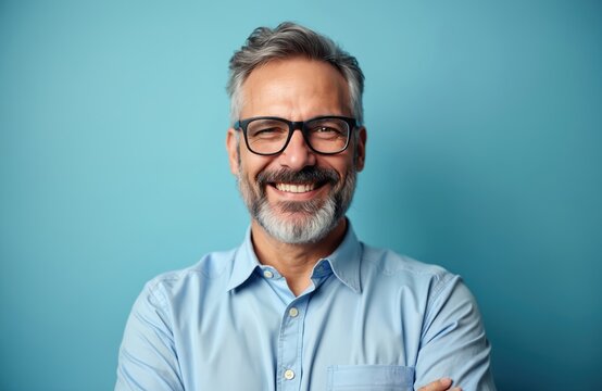 Mature man with gray hair and beard wears glasses and blue shirt, smiles cheerfully against cyan background. Confident pro with friendly expression. Successful adult executive posing for photo.