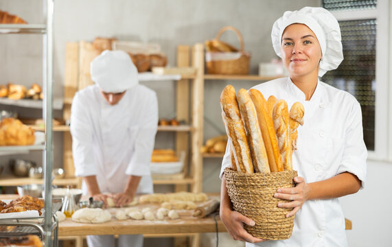Friendly young female baker wearing apron carrying round woven basket with French bread in bakery