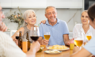 Group of joyful elderly women and man gathered around cozy kitchen table, chatting amiably while drinking beer and wine with snacks. Concept of socialization of older adults