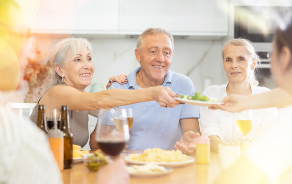 Happy carefree older man and women gathering around kitchen table, sipping alcoholic drinks with snacks, cheerfully chatting, laughing and sharing stories. Seniors communication concept