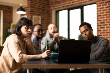 Group of multiethnic friends gathered around table, focused on laptop screen during indoor hangout....