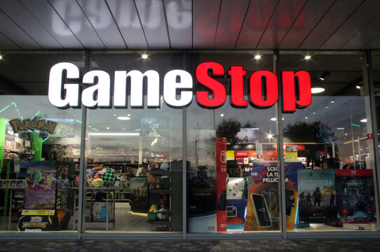 Exterior of a modern GameStop store with large glass windows and bright illuminated sign at night. Facade of an urban gaming shop with reflections and colourful lights.
