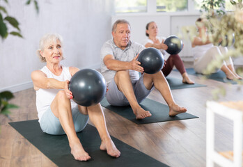 Elderly woman doing pilates with soft ball in group on mat in fitness studio