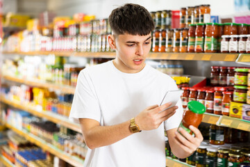 Focused interested young guy making purchases in supermarket, using smartphone to scan barcode on glass jar of canned food. Concept of modern technologies for shopping