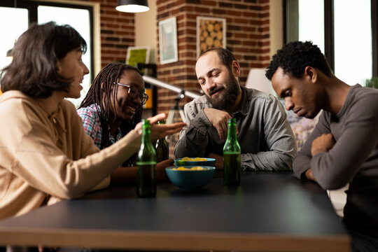 Multicultural friends enjoying casual moment, snacking on chips and sipping drinks while laughing in cozy room. Young group of people laughing and chatting, during weekend hangout.