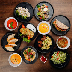 Assorted restaurant dishes flat lay on wooden table with soups salads mains bread and sauces