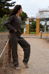 Woman in a brown knitted sweater and wide pants standing near a wooden fence with a green book, looking thoughtfully toward the house on a windy day