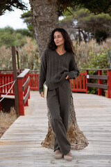 Woman in a dark brown knitted sweater and loose pants leaning against a tree with a book in hand, enjoying a calm autumn day outdoors