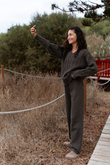 Woman in a dark brown knitted sweater taking a selfie outdoors with a smartphone, standing near dry grass and wooden pathway on a calm autumn day
