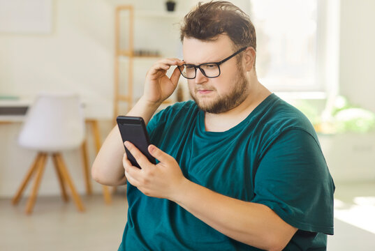 Obese man with glasses reading smartphone screen indoors. Plus-size adult adjusts frames and leans closer at home, struggling to focus on small text. Concept: eyestrain and tech confusion.