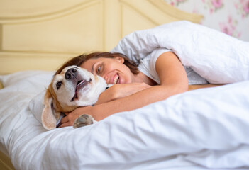 Cozy morning moment with smiling woman hugging her beagle dog under soft blanket in bright bedroom, showing warmth, comfort, companionship and relaxing home atmosphere perfect for pet lovers.
