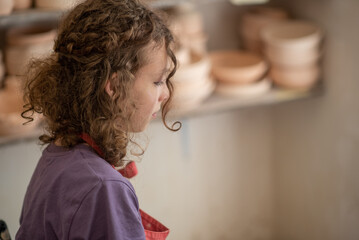 Close-up portrait of a smiling young girl in an apron in a pottery studio. Joyful child surrounded by unfired clay ceramics, emphasizing creative learning and hands-on craft.