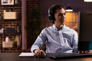 Asian man wearing headphones focusing on computer screen while streaming online webinar. Thoughtful young male adult enjoys learning languages and exploring digital content at home.