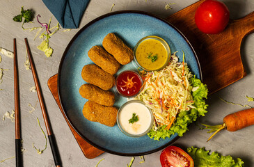 Overhead shot of crispy breaded croquettes served with coleslaw, fresh vegetables, and three assorted dipping sauces on a blue plate. Styled with chopsticks and scattered ingredients