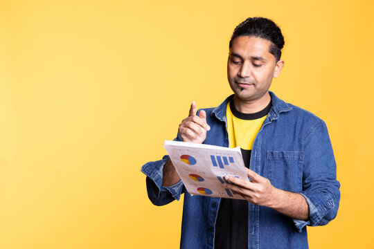 Young male manager looking over company financial profit report audit, reading the results against orange background. Serious indian man reviewing business documents and monthly balance.