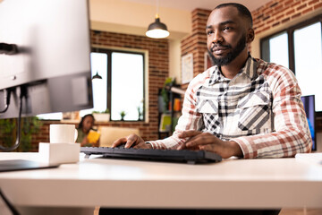 Confident man working from home, checking company revenue and reviewing analytics on computer. Male business advisor uses desktop pc to prepare project presentation in modern brick wall apartment.