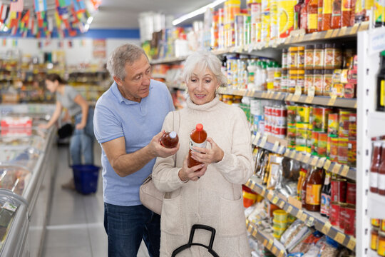 Positive senior couple, man and woman, standing near shelves with canned goods in grocery store, friendly discussing while choosing delicious sauce, examining labels and comparing different bottles