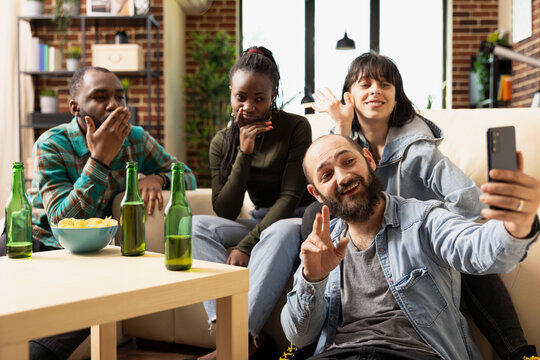 Cheerful diverse friends capturing selfies and sharing weekend hangout at home. Multiethnic group enjoying video calls and mobile interactions, creating memorable moments together.