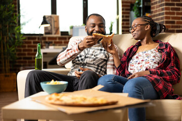 Happy black couple shares fun weekend indoors, watching TV with takeout food and drinks. Boyfriend...