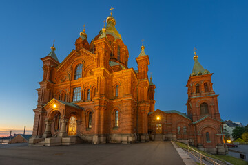 The Uspenski Cathedral, a prominent red-brick Eastern Orthodox cathedral with golden domes, stands illuminated against a clear twilight sky in Helsinki.