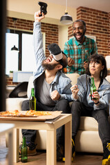 Bearded male gamer wearing VR headset raising wireless controller in excitement after winning 3D gaming tournament. Joyful friends cheering for caucasian man during virtual video game at hangout.
