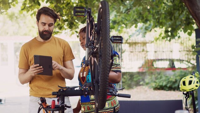 Black woman detaches broken wheel from bike body while caucasian man uses digital tablet to research instructions for mending. Sports-loving couple using smart device for repairing bicycle.