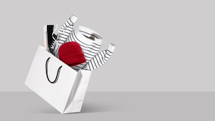 White shopping bag filled with clothing and shoes on soft gray background. Concept of seasonal fashion sale, online shopping, clothing delivery and winter retail, holiday discounts. Copy space. Banner