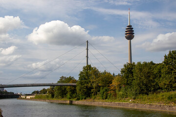 Towering observation structure rises above lush green trees beside a tranquil river, with a modern pedestrian bridge connecting both riverbanks, showcasing urban landscape harmony