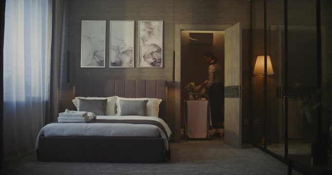 Hotel Maid Entering Elegant Hotel Room With Breakfast Trolley. in Cozy Interior With Stylish Decor, Woman Places Breakfast Cart Next Bed. Guest Care, Professional Hotel Service Routine. Static Shot.