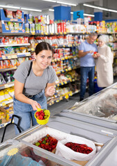 Watchful young woman customer selecting frozen pepper from market fridge in superstore