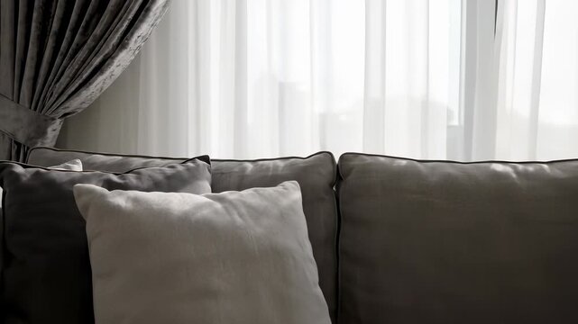 A cozy gray sofa invitingly arranged with multiple pillows in front of a window with soft curtains, evoking feelings of relaxation and serenity, perfect for conveying peacefulness or tranquility.